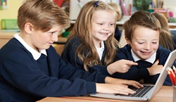Primary school students in uniform gathered around laptop in classroom